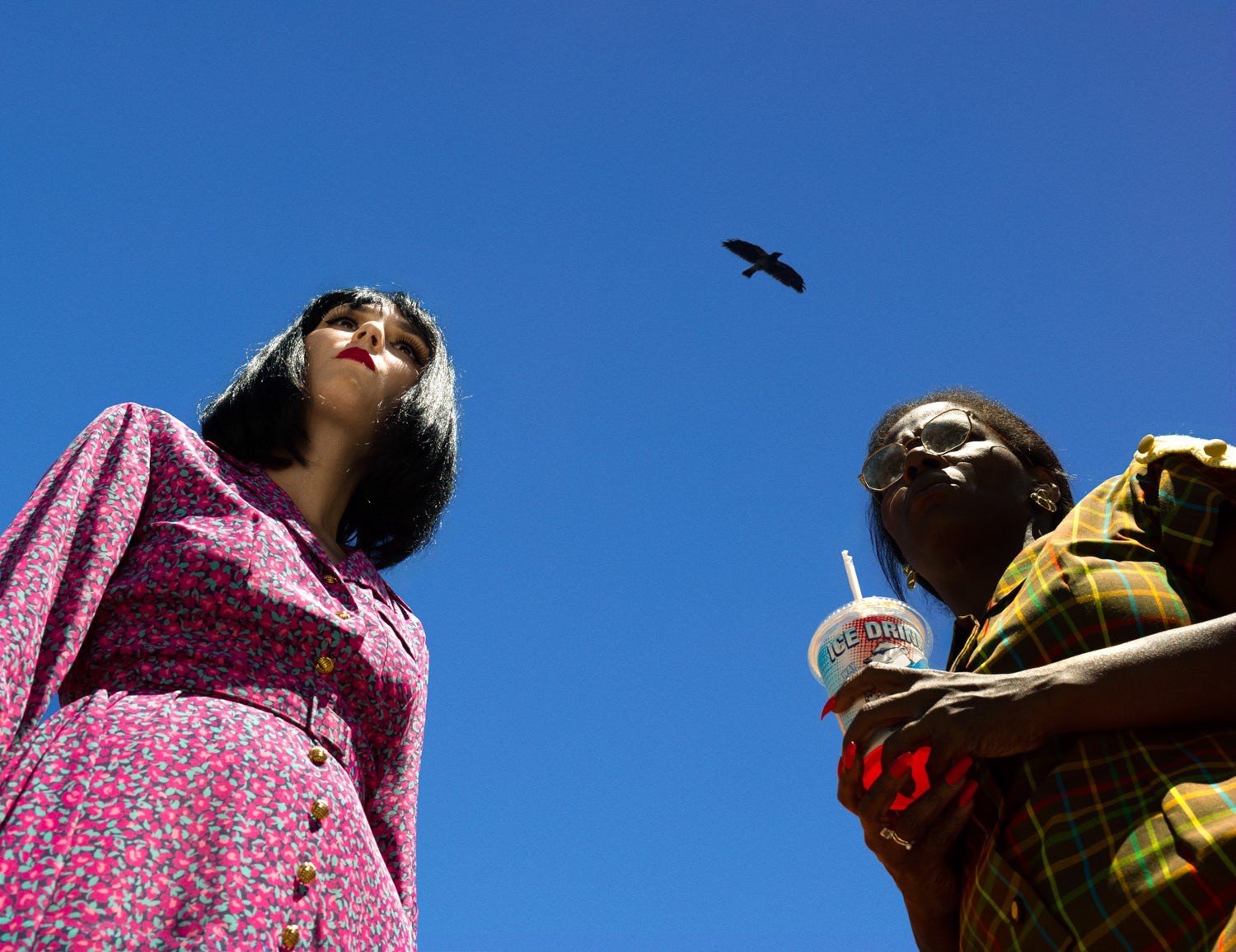 Image by Alex Prager shot from below, featuring one person on the left wearing a floral button-up dress and a bob, next to another person carrying a plastic drink and straw, with a bird flying between them overhead