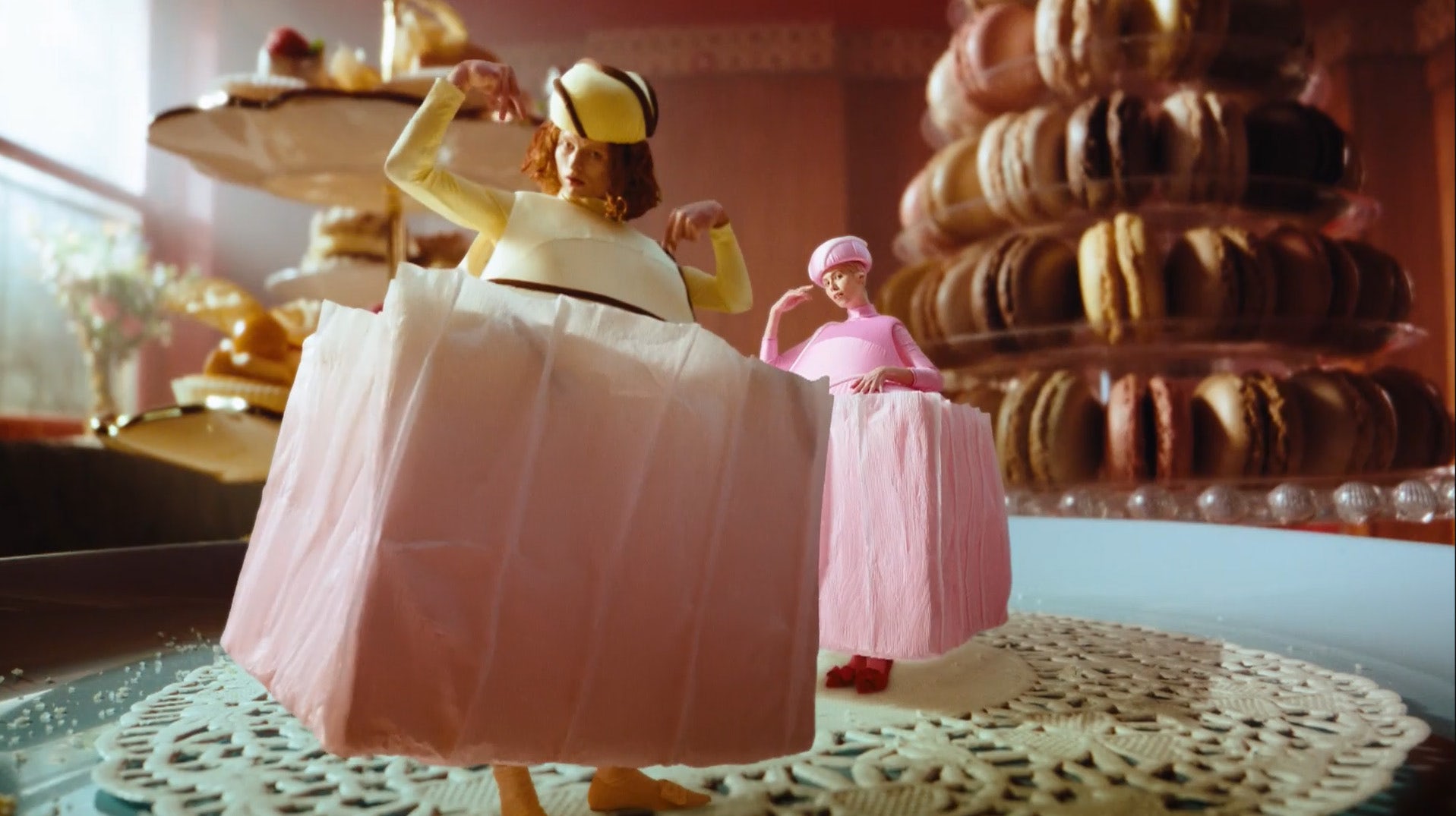 Still from the BBC Eurovision 2023 ad showing two people wearing boxy dresses in the shape of petit-fours with macarons in the background as set dressing