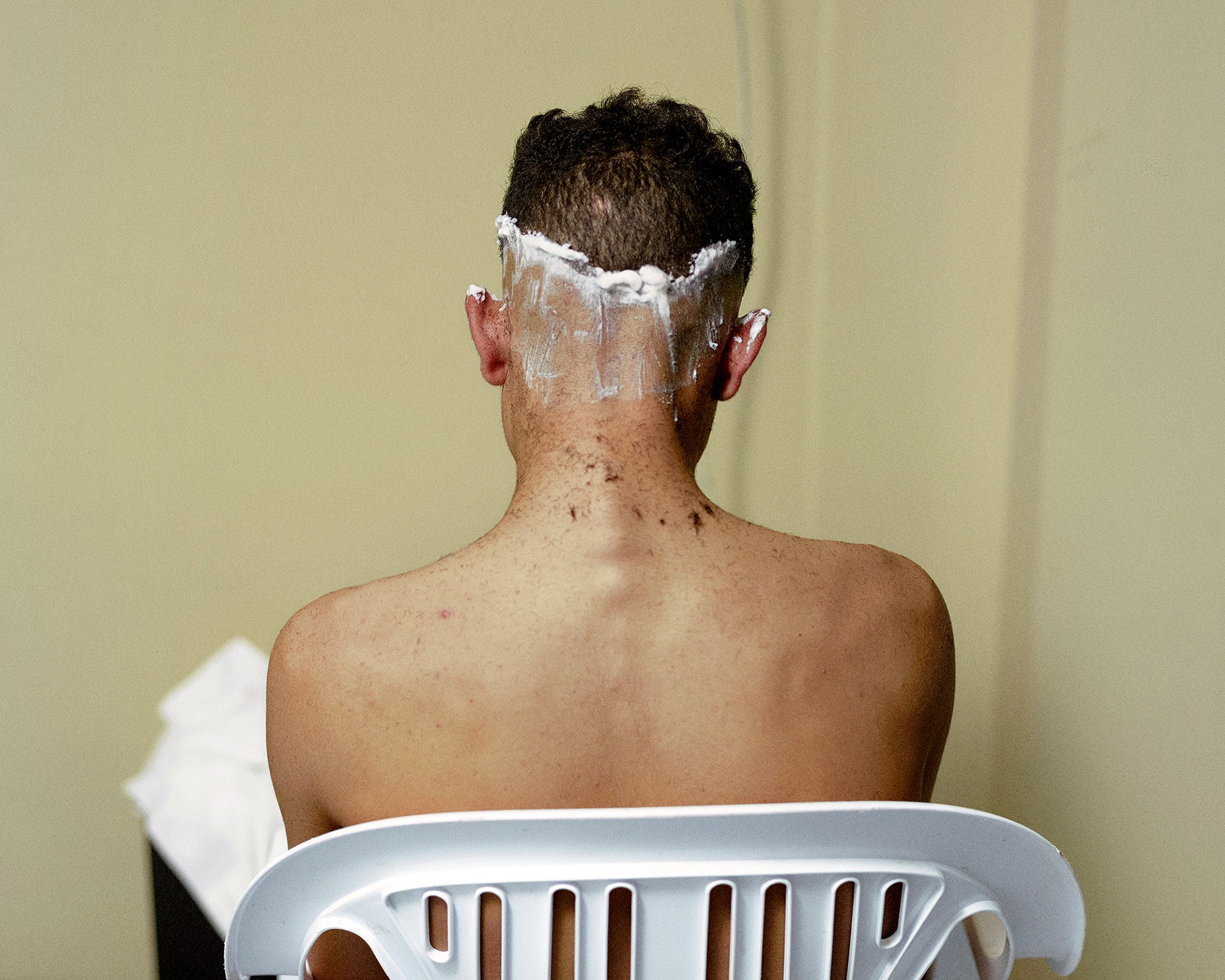 Photograph of a person with shaving cream on the lower half of their head mid-shave, taken from Dialect by Felipe Romero Beltran. The person is sat down in a white chair with their back to the camera