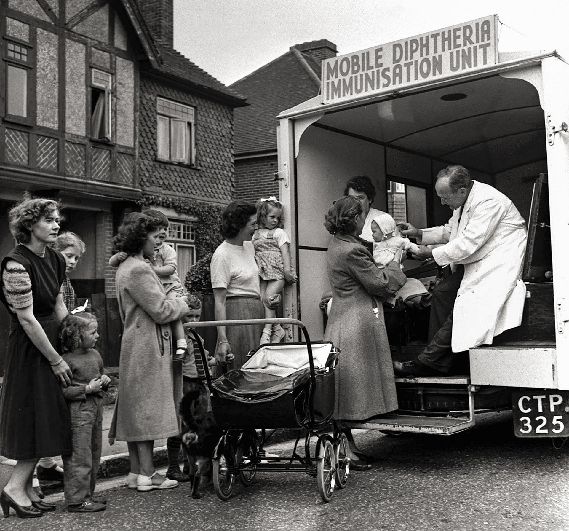 Black and white photograph of people holding children as they queue up for a mobile vaccination centre in the back of a van