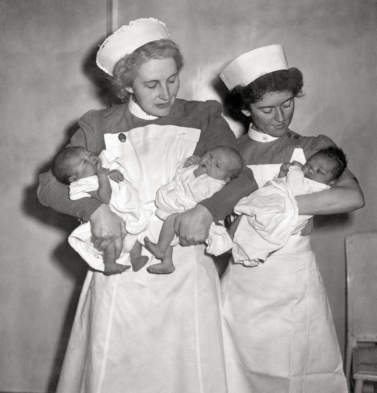 Black and white photograph of two nurses looking down at three babies as they hold them
