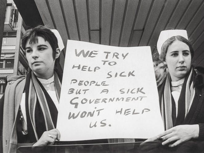 Black and white photograph of two NHS nurses holding up a sign in the middle of them, with the message 'We try to help sick people but a sick government won't help us'