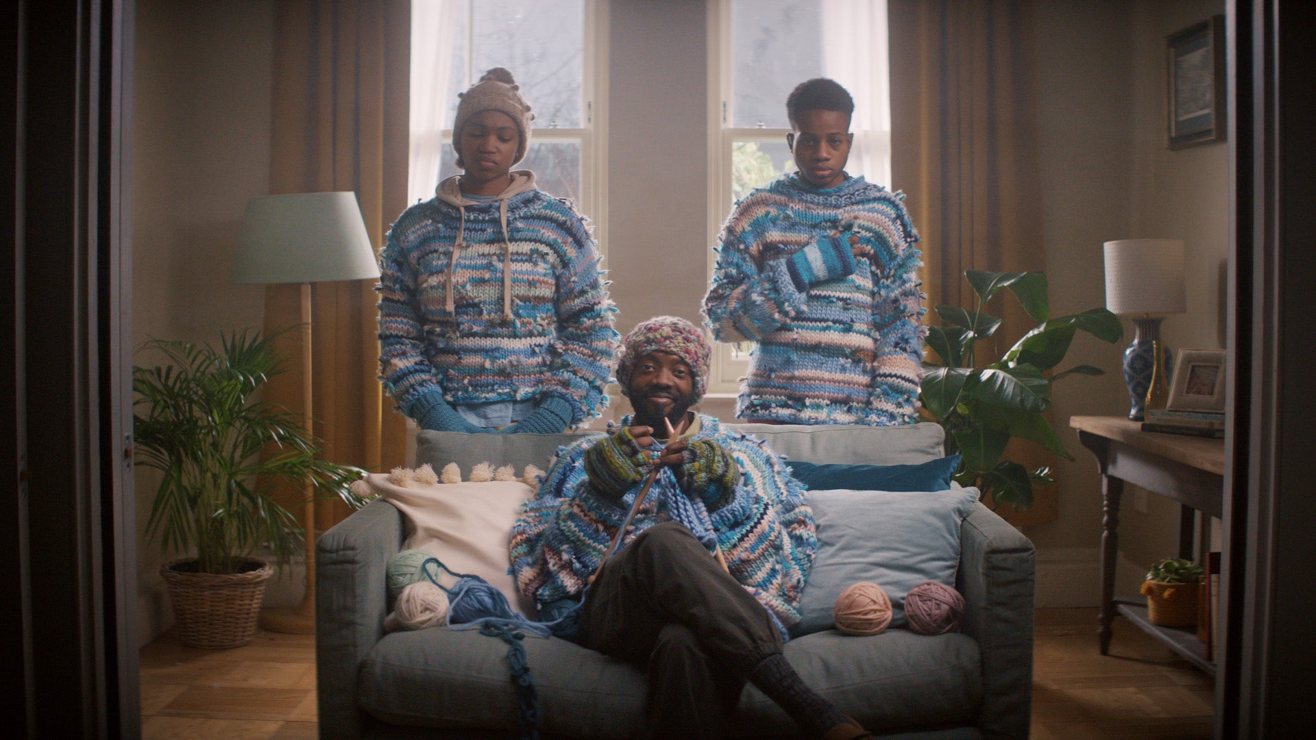 Image of a person sat on a sofa knitting with two people young people stood behind wearing matching knitted patterned jumpers